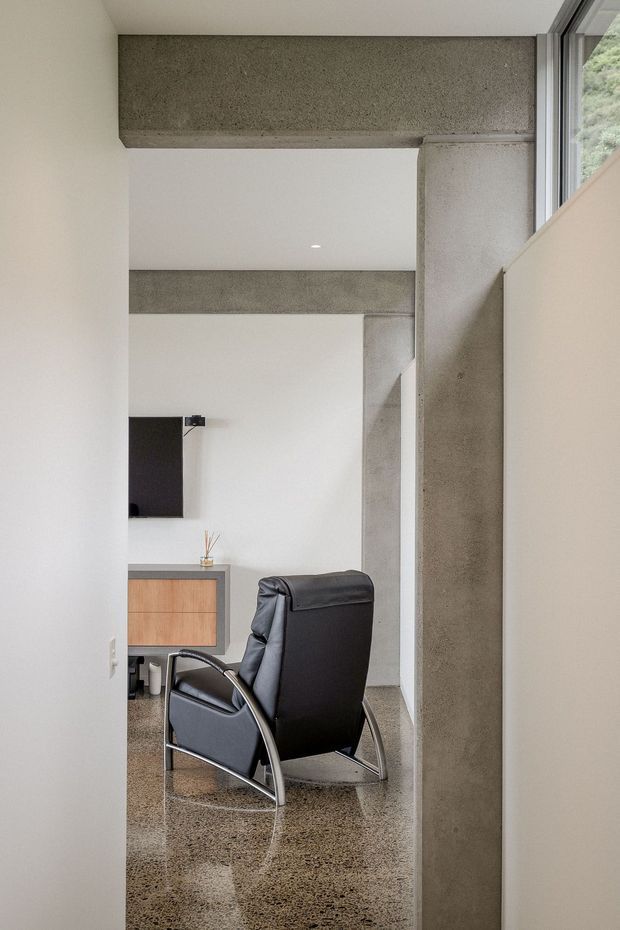 Minimalist interior view through a doorway or passage, featuring exposed concrete structural beams and a concrete column, polished speckled concrete flooring, white walls, and a black leather lounge chair. A timber-fronted built-in cabinet and wall-mounted television are visible in the adjoining space, with a narrow window providing natural light.