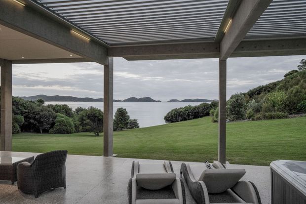 A covered outdoor terrace or pavilion with a louvred roof, substantial concrete columns and beams, and a polished light-grey floor. Outdoor lounge and dining furniture occupy the foreground, opening onto a broad manicured lawn and landscaped coastal outlook with calm water and distant islands. The scene has a restrained contemporary architectural character and an overcast, serene atmosphere.