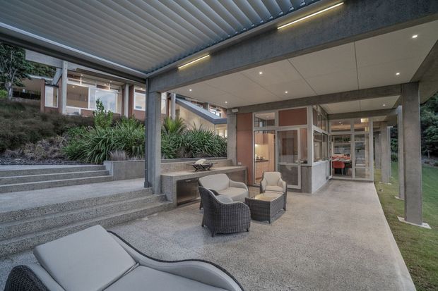 Covered outdoor entertaining area beside a contemporary house, featuring a concrete terrace, outdoor lounge seating, broad steps, integrated planting beds, exposed concrete columns and beams, and a louvred pergola roof. Glazed doors and windows reveal the interior beyond, while landscaped garden surrounds the space.