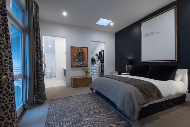 A stylish bedroom with a large upholstered bed, dark navy feature wall, layered grey bedding, patterned area rug, and leopard-print curtains. White walls, recessed lighting, a skylight, and adjoining openings to a bathroom and walk-in wardrobe create a contemporary, refined interior.