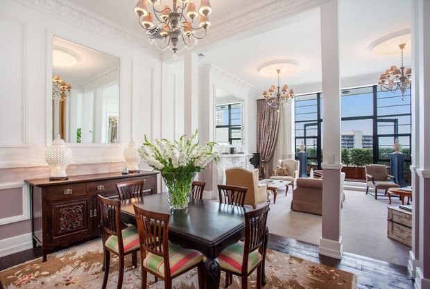 Elegant open-plan dining and living interior with ornate white wall mouldings, tall windows, dark timber dining furniture, a classic sideboard, upholstered seating, and statement crystal chandeliers. The room has a refined traditional character with abundant natural light and views of an urban setting through the glazing.