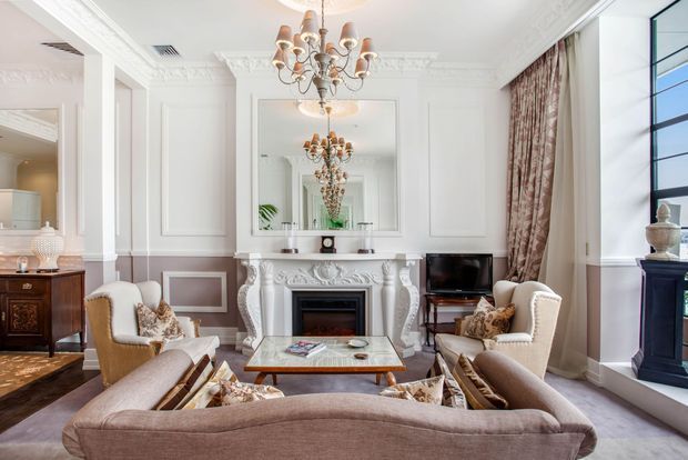 Elegant formal living room with ornate white wall mouldings, a carved decorative fireplace mantel, large mirror, chandelier, tall curtained windows, and upholstered seating arranged around a central coffee table. The room uses a refined neutral palette with taupe and blush upholstery, white architectural detailing, and dark timber accent furniture.
