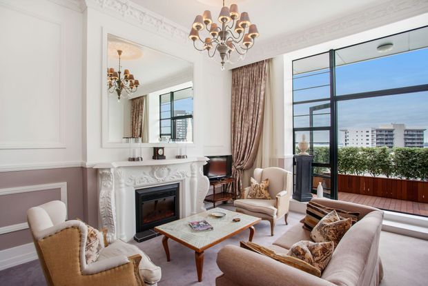Elegant classical living room with ornate white decorative mouldings, a carved fireplace surround, large overmantel mirror, chandelier, and soft taupe wall panelling. A seating arrangement surrounds a wooden coffee table, while full-height black-framed glazing opens to a balcony or terrace with an urban view.