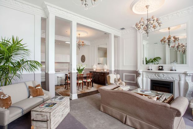 Elegant, bright formal living room opening onto a dining area, with ornate white wall panelling, decorative ceiling mouldings, mirrored surfaces, a carved white fireplace, chandeliers, upholstered seating and a large indoor palm. The room has a refined traditional character with soft neutral furnishings and grey-beige tones.