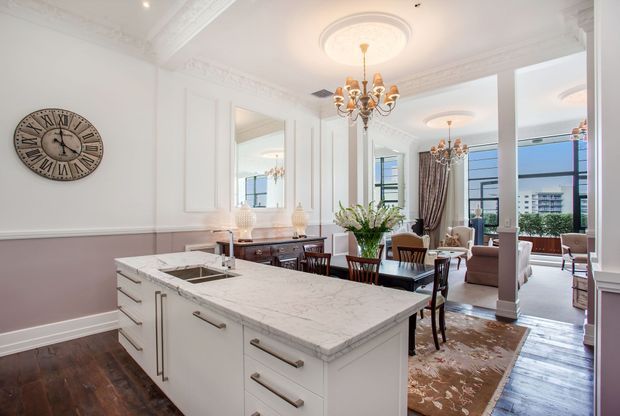 Bright, elegant open-plan kitchen and dining/living area with a large white marble island, white cabinetry, dark timber flooring, pale mauve wall panelling, ornate ceiling mouldings, decorative ceiling medallions and chandeliers. A dining table, lounge seating, tall mirrors, and large windows are visible beyond the island.