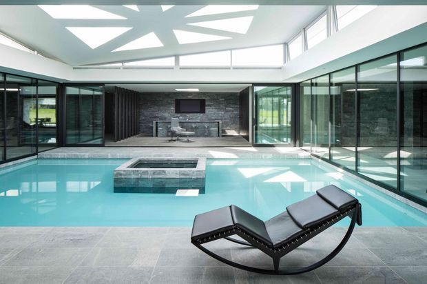 A bright, expansive indoor pool area with turquoise water, a raised rectangular spa, and a black sculptural chaise lounge in the foreground. Floor-to-ceiling black-framed glazing surrounds the pool, with a visible living or media area beyond. The space features a dramatic geometric skylight and a dark stone feature wall, creating a sleek contemporary architectural interior.