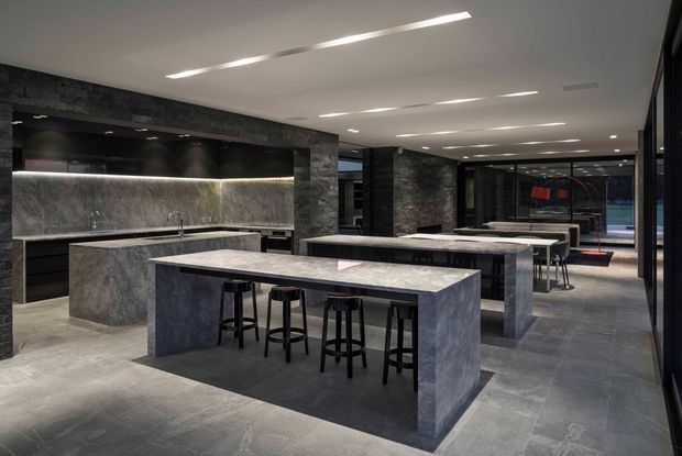 A large, sophisticated communal kitchen and entertaining space with multiple stone-clad islands, integrated dark cabinetry, bar stools, long dining or serving tables, and dramatic charcoal stone walls. Linear recessed ceiling lighting and full-height glazing create a contemporary, refined atmosphere.