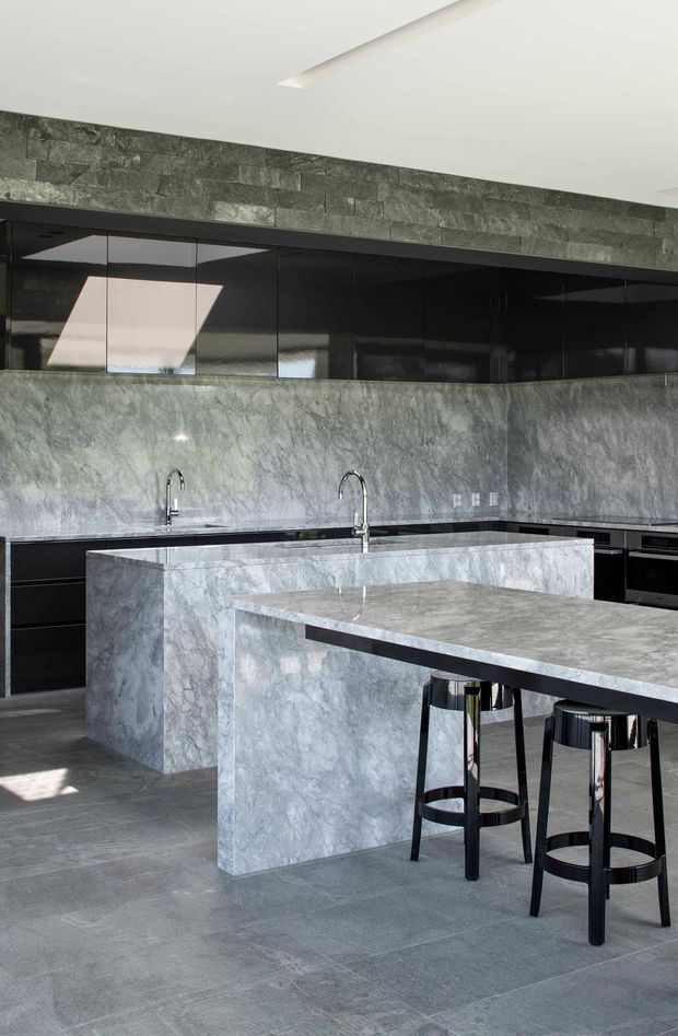 Contemporary kitchen featuring extensive grey veined stone cladding across the island, benchtops and splashback, with sleek glossy black cabinetry, integrated appliances, dark tiled flooring, and a polished chrome island stool arrangement. A narrow reflective window band and recessed ceiling lighting add a streamlined, dramatic character.