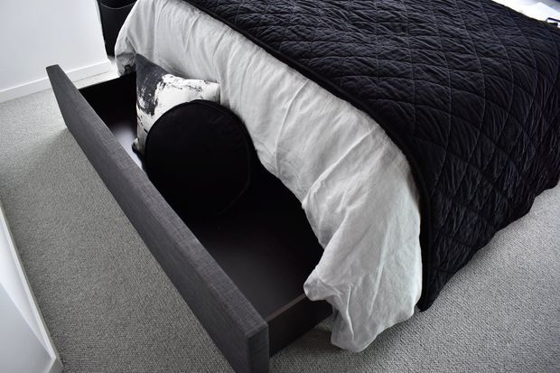 Close-up view of a contemporary bedroom with a bed featuring a dark textured upholstered frame and black quilted coverlet over white bedding. A large under-bed storage drawer is open, revealing integrated storage. The room has light grey carpet, white walls, and a simple monochrome palette.