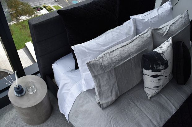 Close-up view of a contemporary bedroom corner with a neatly made bed layered in white and grey linen bedding, charcoal and patterned cushions, and a dark upholstered headboard. A round concrete side table with a small lamp and glass sits beside the bed, next to large black-framed glazing overlooking an outdoor area.