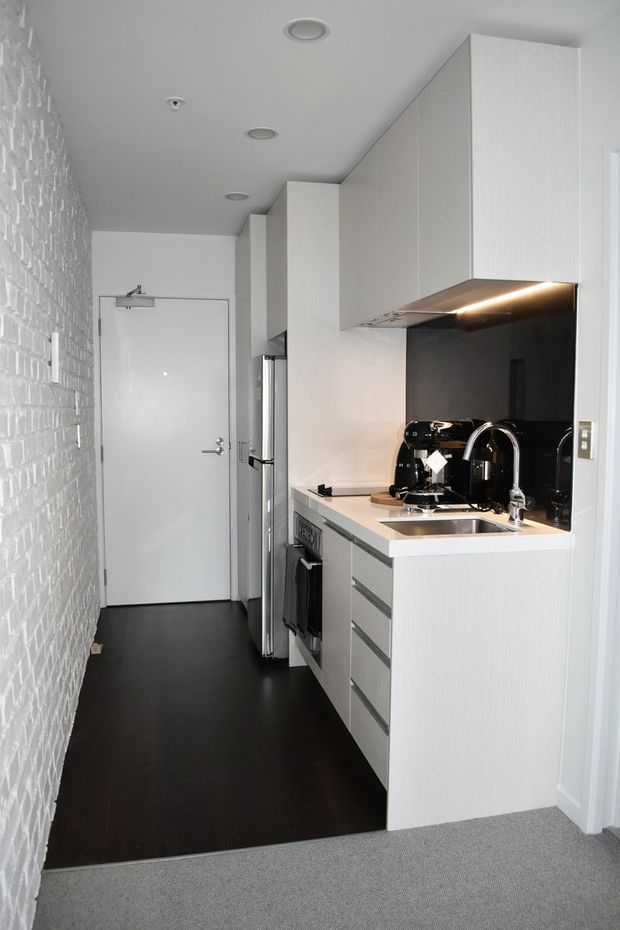 A compact, narrow galley kitchen with white vertical-grain cabinetry, integrated appliances, a stainless-steel sink and tap, and a glossy black splashback beneath illuminated overhead cabinets. The kitchen opens from a carpeted area, with a dark hard floor, white painted brick feature wall, recessed ceiling lights, and a white entry door at the rear.