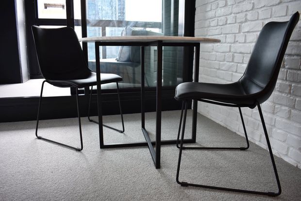 A compact, minimalist dining or meeting nook with a round timber tabletop on a black metal pedestal frame, paired with two black molded chairs. The space features a white-painted exposed brick wall, dark-framed floor-to-ceiling glazing, and a light grey carpet, creating a restrained industrial-modern atmosphere.