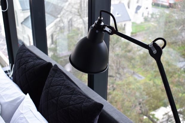 Close-up interior view of a black articulated task lamp positioned beside a bed, with black quilted pillows and white bedding in the foreground. Floor-to-ceiling dark-framed glazing provides a bright urban or residential outlook beyond.
