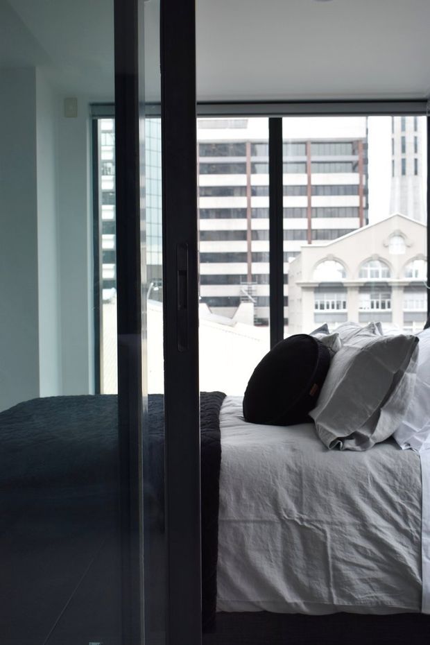 A contemporary urban bedroom viewed through a dark-framed glass sliding door. The room features a neatly made bed with white bedding, a dark round cushion and dark throw, pale walls, and large windows overlooking dense city buildings. The space has a quiet, minimal and slightly moody atmosphere.