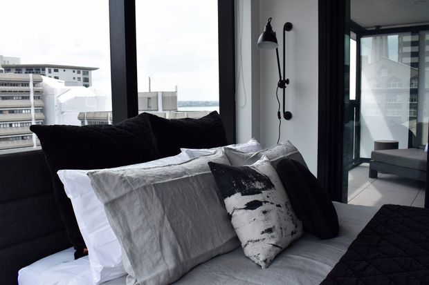 A contemporary bedroom viewed at close range, featuring layered white and grey bedding, large black cushions, and a monochrome patterned accent pillow. Floor-to-ceiling black-framed glazing provides expansive urban and coastal views, while a black wall-mounted adjustable light adds an industrial touch. A glimpse of an adjoining living area is visible through the glazed opening.