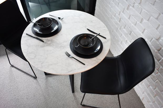 A compact dining area with a round white marble-look table set for two, featuring black plates, bowls and cutlery. Two sculptural black dining chairs sit beside a white-painted exposed brick wall, with dark-framed glazing visible behind.
