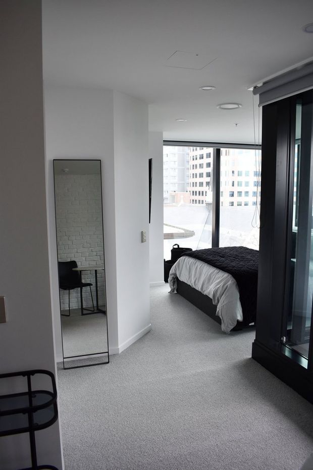 A minimalist bedroom in a contemporary apartment, viewed from an adjoining area. The room has a low dark bed with white bedding and a dark quilt, wall-to-wall light grey carpet, white walls and ceiling, expansive floor-to-ceiling glazing with dark frames, and roller blinds. A full-length black-framed mirror reflects a small black desk and chair. High-rise buildings are visible outside.