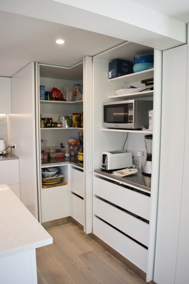 A compact, highly organised kitchen pantry and appliance-storage area with full-height white cabinetry, open shelving, drawers, and integrated countertop appliances including a microwave, toaster and blender. A light stone-look kitchen benchtop and pale timber-look flooring are visible in the foreground and floor area. The space is brightly lit and efficiently planned.