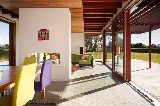 Bright open-plan modern living and dining space with extensive floor-to-ceiling glazing and large glazed sliding doors opening onto a covered terrace and lawn. The room features a polished or honed concrete floor, exposed dark timber ceiling beams, timber slatted ceiling sections, colourful dining chairs, and a green sofa. Strong indoor-outdoor connection and abundant natural light define the space.
