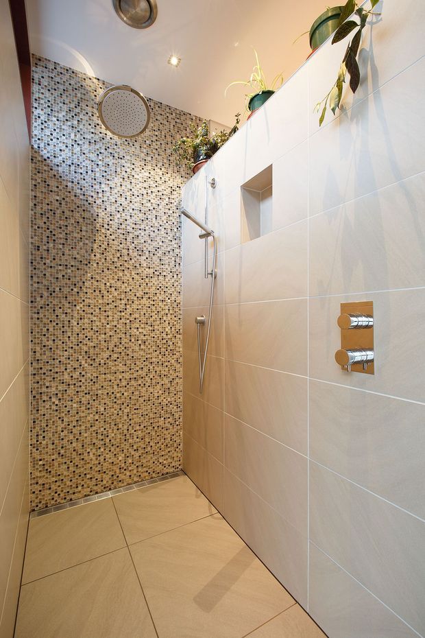 A compact modern walk-in shower with large beige wall and floor tiles, a contrasting mosaic-tile feature wall, recessed shelf, chrome shower fittings, and potted plants above the wall partition. The space is brightly lit with clean contemporary detailing.