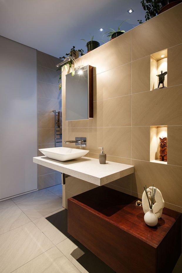 A contemporary bathroom vanity area with a wall-mounted white vessel basin, floating light benchtop, dark timber cabinetry, large beige wall tiles, recessed display niches, a mirrored cabinet, and integrated accent lighting. Potted plants and decorative objects add warmth to the refined space.