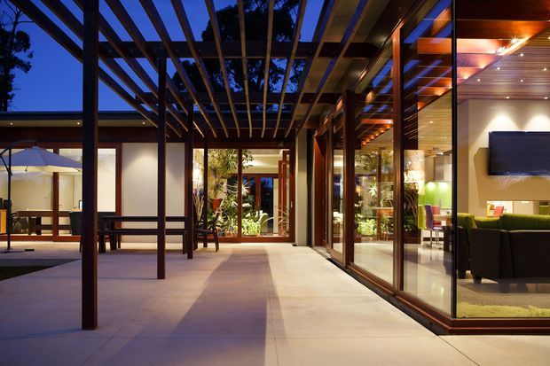 A contemporary residential courtyard or covered outdoor living area viewed at dusk, featuring a timber pergola with exposed beams, large glazed sliding doors, tiled paving, and warm interior lighting. The glazing reveals an adjacent living room with a sofa, television, and rug, while a landscaped courtyard and outdoor seating extend beyond.