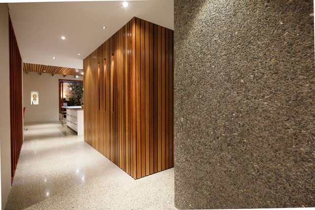 A refined contemporary interior hallway featuring a polished light terrazzo floor, warm vertical timber slat wall panelling, and a textured dark stone or concrete feature wall. Recessed ceiling lighting illuminates a partially visible kitchen or service area at the end of the corridor, while timber slats continue across the ceiling in the background.