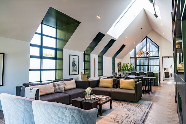 Bright, spacious contemporary open-plan living room with a large sectional sofa, armchairs, coffee tables and rug, extending into a dining area and kitchen. The room features dramatic angled ceilings, extensive black-framed glazing, skylights, dark green structural or feature panels, and a tall glazed gable-like end wall.