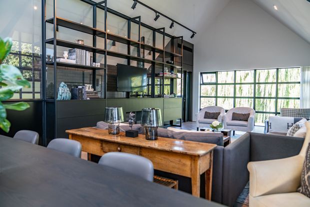 A sophisticated open-plan living and dining area with a large black-framed steel shelving and cabinetry wall, integrated television, exposed display shelving, and extensive glazing. A rustic timber console table contrasts with contemporary upholstered seating, while track lighting and a high sloped ceiling add architectural interest.