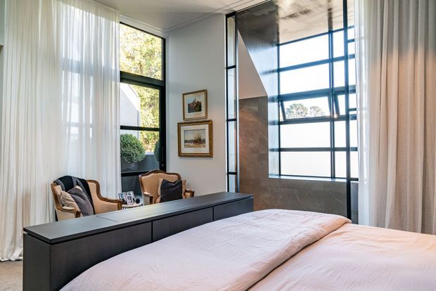 A refined bedroom with a large upholstered bed in the foreground, dark built-in cabinetry or a low partition behind it, and two classic upholstered armchairs in a sitting area by tall windows. Sheer full-height curtains soften abundant natural light, while a dramatic black-framed glazed architectural feature and stone-toned wall create a strong focal point. Framed artwork and neutral bedding complete the calm, elegant interior.