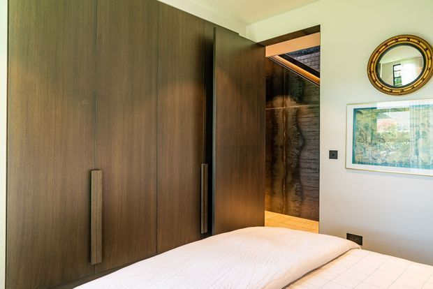 A warm, contemporary bedroom viewed toward a large built-in dark timber wardrobe with long vertical handles. The bed occupies the foreground, while an open doorway reveals a darker textured feature wall and warm-lit adjoining area. White walls, framed artwork, a round decorative mirror, and integrated dark electrical fittings complete the scene.