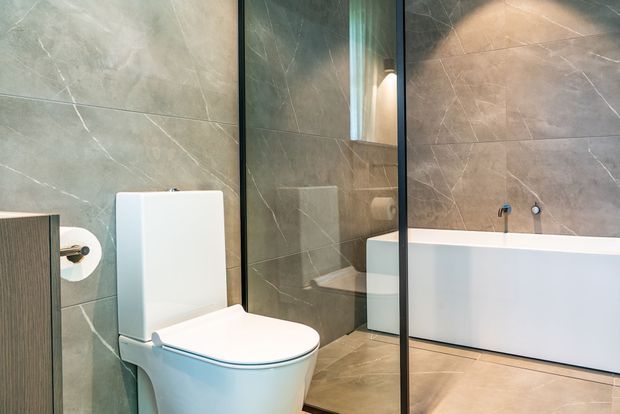 Contemporary bathroom featuring large-format grey stone-look wall and floor tiles, a wall-adjacent white toilet, a freestanding white bathtub, and a frameless glass shower partition. Dark vertical framing and warm integrated lighting add contrast to the otherwise neutral, refined space.