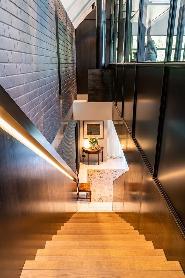 A dramatic interior stairway descends toward a small entry or living area. The stairs have warm timber treads, with dark timber wall panelling and integrated linear lighting. A pale painted brick wall runs along the left side, while black-framed glazing and dark cabinetry or wall panels line the right. At the bottom is a console table with flowers, artwork, a patterned rug, and glimpses of bright curtains and an adjacent room. The space has strong vertical lines, contrasting materials, and abundant natural light from the upper glazing.