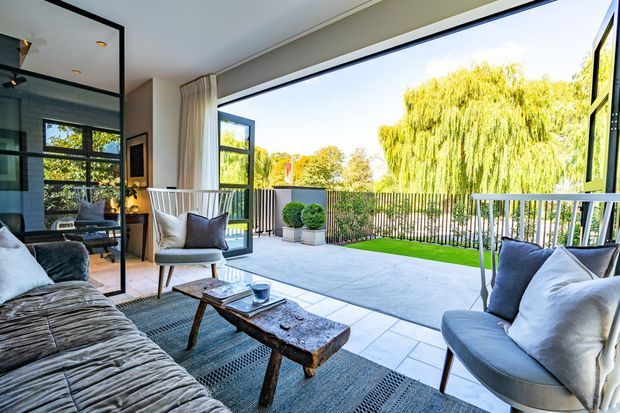A bright contemporary living room opening onto a spacious paved terrace through expansive black-framed glazing and folding doors. The room features a large textured sectional sofa, upholstered lounge chairs, a rustic timber coffee table, patterned rug, neutral cushions, and minimal decor. A balcony-style railing and lush weeping tree are visible beyond the terrace.