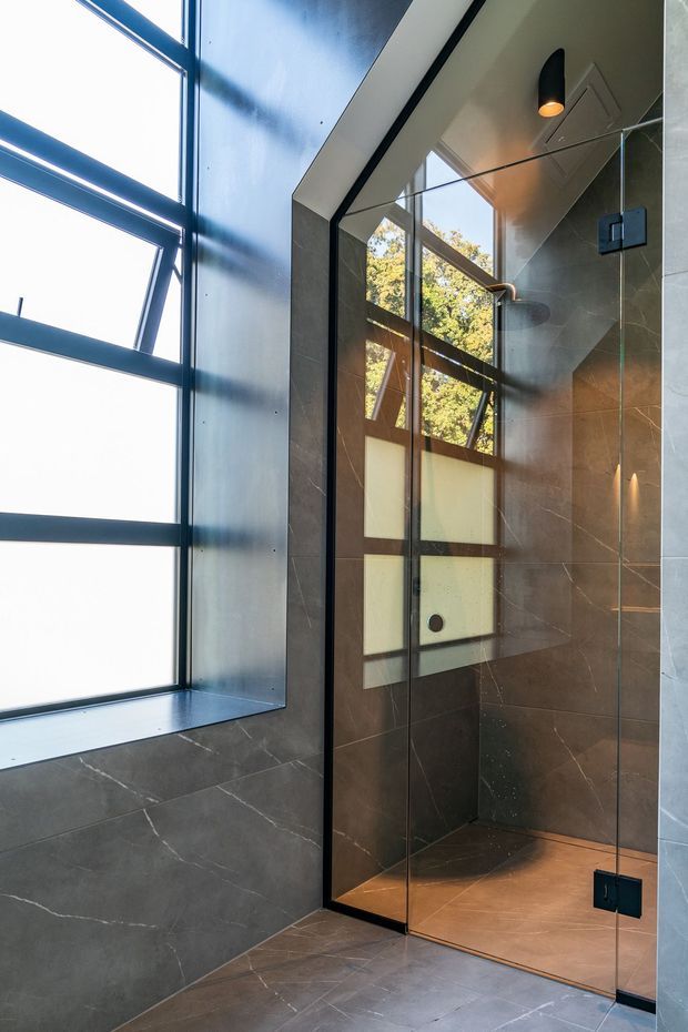 Contemporary walk-in shower enclosed by a frameless glass screen, with large dark grey stone-look wall and floor tiles, black hardware, a wall-mounted showerhead, and a tall angular window providing natural light.