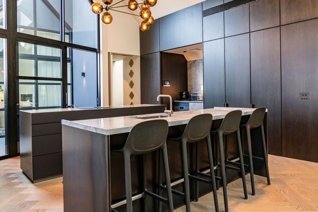 A sophisticated contemporary kitchen with a large central island, pale stone benchtop, dark timber-look cabinetry, integrated appliances, and four bar stools. Floor-to-ceiling glazing brings in natural light, while a sculptural cluster pendant adds a warm decorative accent. The timber floor is laid in a herringbone pattern.