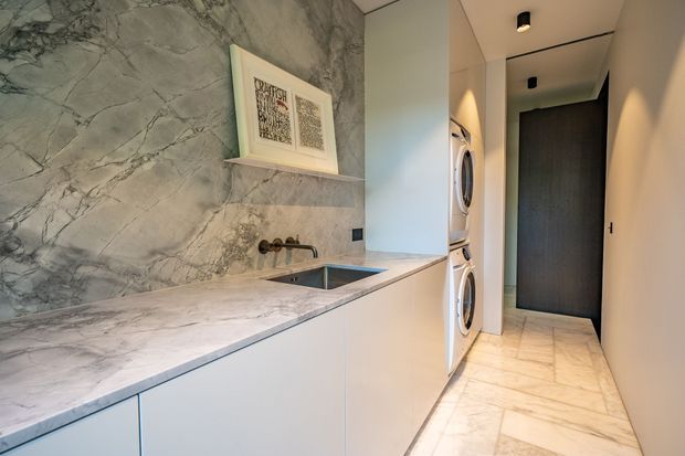 A narrow, contemporary laundry room with floor-to-ceiling marble-look wall cladding behind a long white built-in counter. The counter incorporates a small undermount sink and wall-mounted tapware, with front-loading washer and dryer stacked in a recess. Pale stone-look flooring continues down the hallway toward a dark timber-toned door, while recessed and black ceiling lights provide subtle illumination.