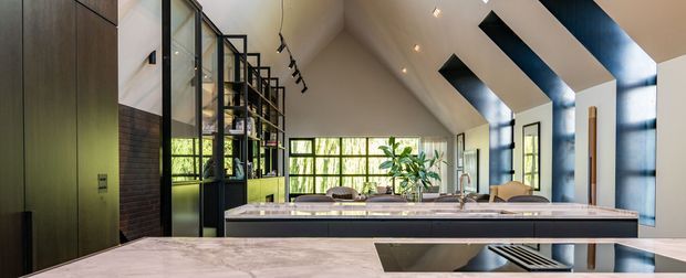 A contemporary open-plan kitchen and living space with a dramatic vaulted ceiling, exposed dark ceiling beams, clerestory-style glazing, and extensive black-framed windows. The kitchen features a long stone island with an integrated sink, dark built-in cabinetry, and a black-framed glass display partition. A dining or living area and greenery are visible beyond the island.