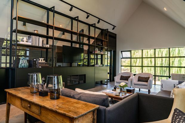 A sophisticated contemporary living room with a high pitched ceiling, expansive black-framed glazing, and a dramatic floor-to-ceiling open shelving and cabinetry wall. Dark built-in joinery incorporates a television and fireplace, while neutral upholstered seating surrounds a low coffee table. A substantial rustic timber console table with metal accents adds warmth against the darker palette.