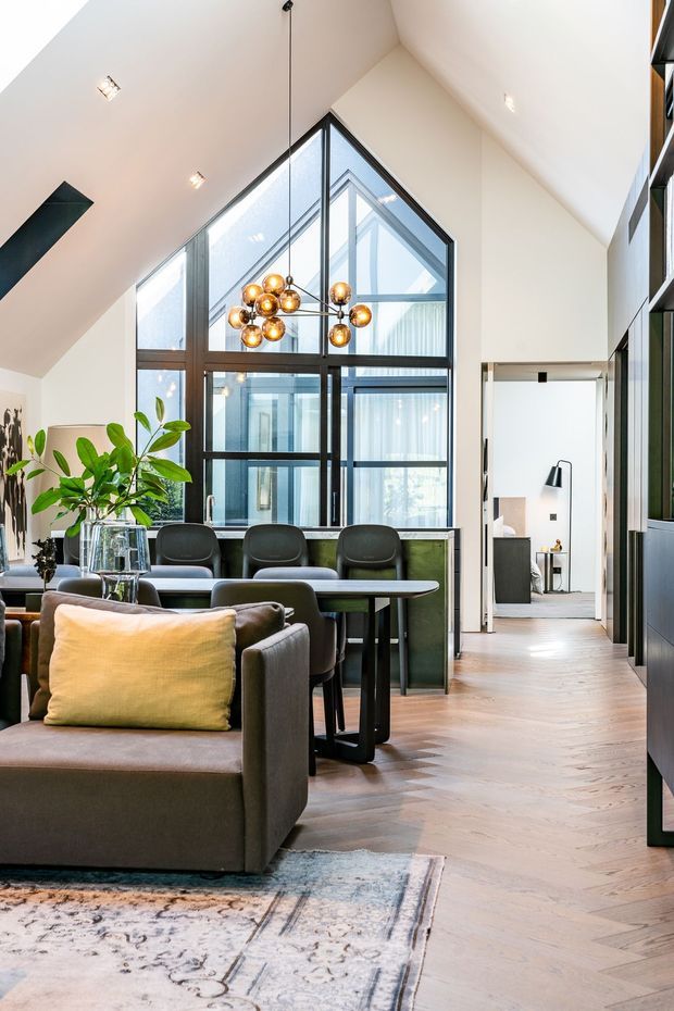 A sophisticated open-plan living and dining area with a dramatic pitched ceiling and full-height black-framed glazing. A sculptural cluster pendant hangs above the dining table, while a large grey sofa, dining chairs, indoor plants, and a patterned rug create a refined contemporary interior. Pale herringbone timber flooring contrasts with dark cabinetry and window frames. An adjoining room is visible through a doorway.