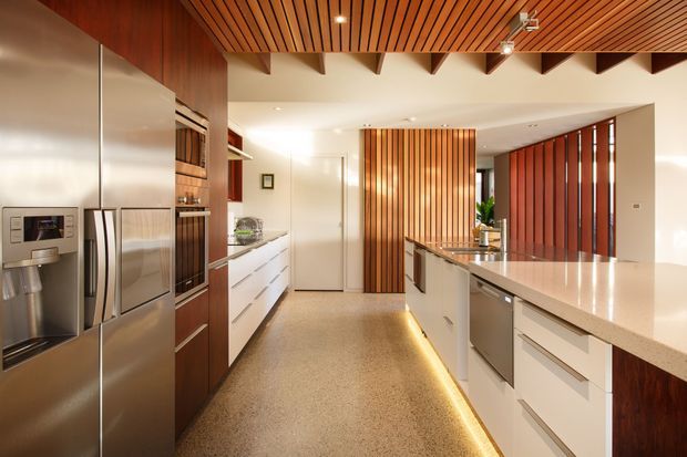 A warm contemporary kitchen viewed from the main workspace, featuring a large island with pale stone benchtop, white drawers, dark timber cabinetry, integrated stainless-steel appliances, a polished concrete or terrazzo-like floor, and extensive timber slatted ceiling and screening. Recessed and under-cabinet lighting create a refined, inviting atmosphere.