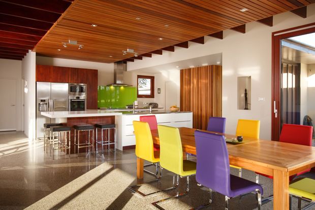 Bright open-plan kitchen and dining area with a vivid green splashback, white island cabinetry, stainless-steel appliances, and a large timber dining table surrounded by colourful modern chairs. The room features a prominent warm timber slatted ceiling with exposed dark beams, polished dark flooring, timber wall accents, and large glazed doors opening to an outdoor area.