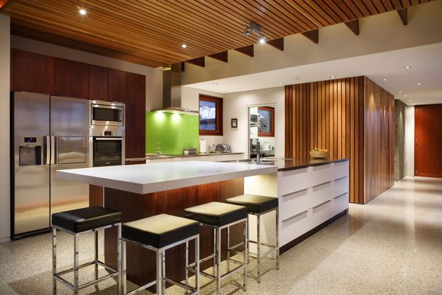 A spacious contemporary kitchen with a large white island and four black bar stools. Dark timber cabinetry, stainless-steel appliances, a vivid green splashback, and extensive timber slatted ceiling and wall detailing create a warm, modern interior. The open-plan space includes a bright polished floor and visible structural ceiling beams.