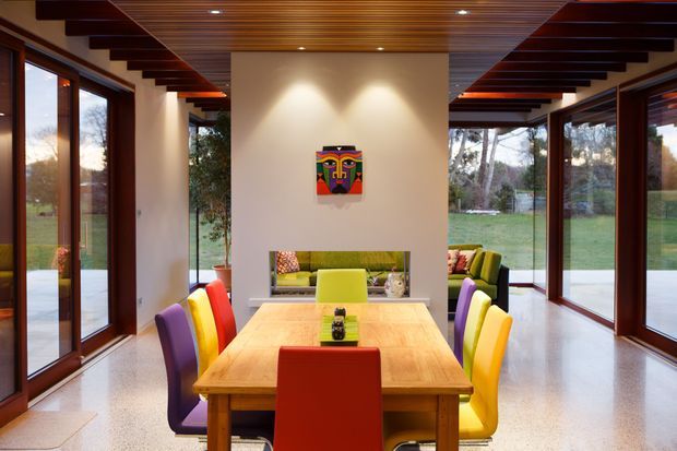 A bright, modern dining room centered on a large timber dining table with vividly upholstered red, yellow, purple, and lime-green chairs. Full-height timber-framed glazing wraps both sides of the room, opening views to the garden. The room features a warm timber slatted ceiling with exposed dark timber beams, polished light terrazzo flooring, white walls, and a colourful artwork above a rear built-in or reflected seating area.
