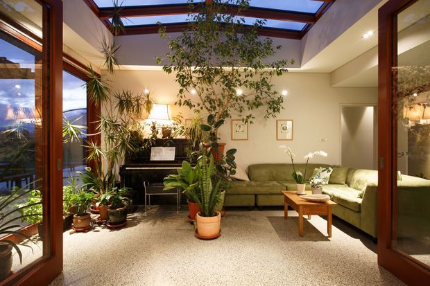 A warm, plant-filled sunroom or living area with extensive timber-framed glazing and a series of overhead skylights. The room features a green sectional sofa, upright piano, coffee table, indoor plants, framed artwork, and a speckled terrazzo-style floor. Views through the glazing show an exterior balcony and distant landscape. The space feels bright, leafy, relaxed, and connected to the outdoors.