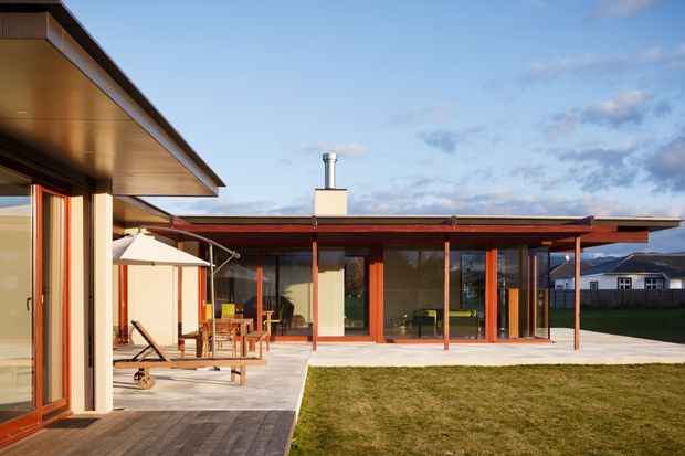 Single-storey modern residential courtyard composition with broad low-pitched roof planes, extensive floor-to-ceiling glazing, warm timber framing, and a paved patio opening onto a lawn. Outdoor dining and lounge furniture sit beneath the covered terrace, while the glazed living area and adjacent house wings frame the view. The image is brightly lit with a restrained palette of timber, dark grey, off-white, and green.
