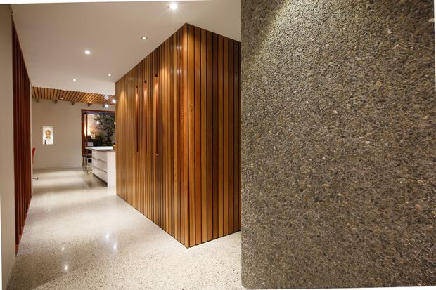 A refined residential hallway with polished light terrazzo flooring, warm vertical timber battens forming a feature wall and ceiling detail, recessed lighting, and a textured dark stone or concrete feature wall in the foreground. The corridor leads toward a bright kitchen or living area with white cabinetry visible in the distance.