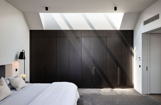 A bright, contemporary bedroom with a low-profile bed in the foreground and a full wall of dark timber built-in wardrobes. A large linear skylight casts strong diagonal beams of daylight across the cabinetry and carpeted floor. The room has white walls and ceiling, minimalist black ceiling spotlights, a black floor lamp, and a simple grey-framed doorway.
