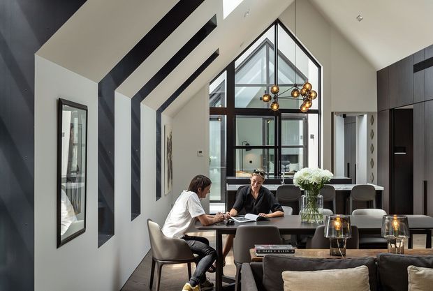 A sophisticated open-plan interior combining a dining area, living room, and kitchen. The space features a dramatic vaulted ceiling with bold black linear accents, floor-to-ceiling black-framed glazing, dark built-in cabinetry, a dining table with upholstered chairs, and a contemporary sculptural pendant light. Two people are seated at the dining table, while a sofa and coffee table occupy the foreground.