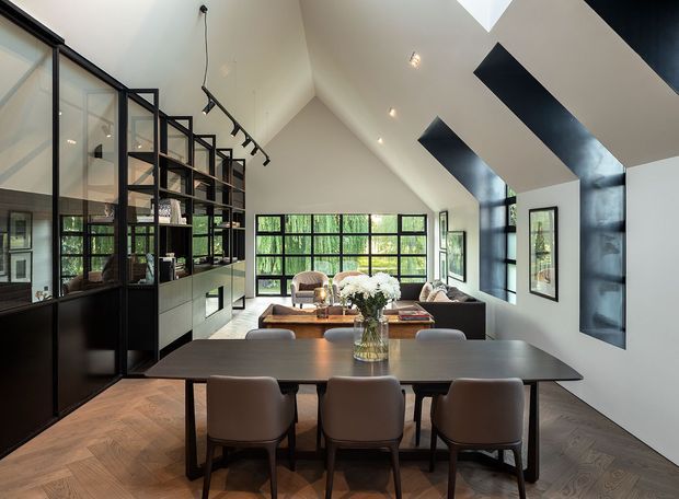 Open-plan dining and living area with a dramatic vaulted ceiling, extensive black-framed glazing, and a full-height black metal and glass shelving partition. A long dark dining table with upholstered chairs occupies the foreground, while a sectional sofa and coffee table sit beyond. White walls and ceiling contrast with black architectural accents, skylights, and warm herringbone timber flooring.